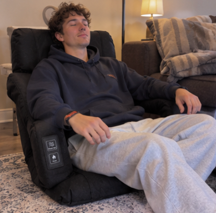 A man reclining comfortably in a black electric floor chair with a control panel on the armrest, set in a cozy living room environment.